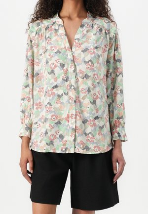 Femme portant un chemisier à boutons fleuri pastel à manches longues, associé à un short noir arrivant aux genoux.