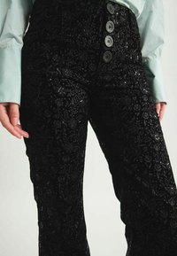 Pantalon noir brodé avec des motifs floraux, orné de paillettes, et une taille haute avec des boutons décoratifs. Texture lisse.