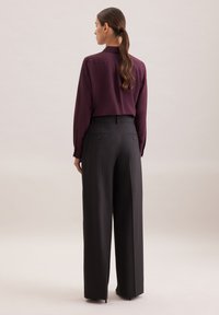 Bordeauxfarbene Bluse mit langen Ärmeln, kombiniert mit hoch taillierten, schwarzen, weit geschnittenen Hosen, die über Rückentaschen und einen glatten, strukturierten Stoff verfügen.