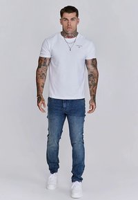 Balta īsroku T-krekls ar smalku logotipu, kopā ar zilām džinsa biksēm un baltām sporta kurpēm. Modelis stāv taisni pret neitrālu fonu.