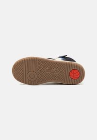 Sneaker sole in brown rubber with a textured grip pattern, featuring a circular logo in red and a basketball design at the heel.