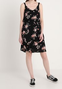 Musta kukallinen mekko, jossa on vaaleanpunaisia ja valkoisia kukkia, V-aukko, kapeat hihnat, gathered vyötärö ja lentävä hame. Yhdistettynä mustiin lenkkareihin.