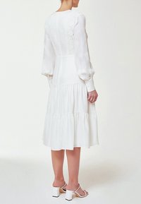 Biała długa sukienka z długim rękawem i warstwową spódnicą, z ozdobnym tyłem z guzikami i kwiatowymi akcentami. Gładki materiał o lekko przezroczystym wykończeniu.