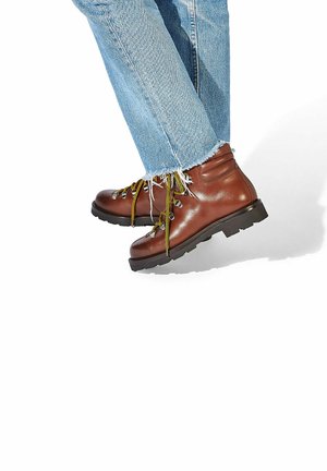 Brązowe skórzane buty trekkingowe z czarnymi gumowymi podeszwami, z żółtymi sznurówkami i srebrnymi oczkami, w zestawieniu z zniszczonymi niebieskimi dżinsami.