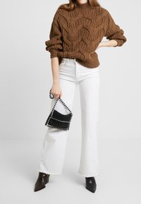 Bruine gebreide trui met een golvende textuur, gecombineerd met witte flared jeans. Zwarte petten handtas en puntige zwarte enkellaarsjes als accenten.