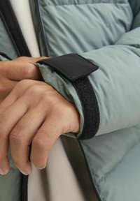 Padded jacket in light green with black Velcro cuff. Features visible stitching and a smooth, shiny texture on fabric.