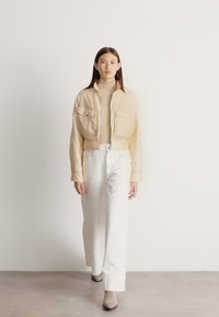 Sieviete pastaigājas iekštelpās, valkājot bēša krāsas īsu žaketi, bēša krāsas augstu apkakli, baltas platām kājām bikses un bēša krāsas potīšu zābakus uz vienkārša fona.