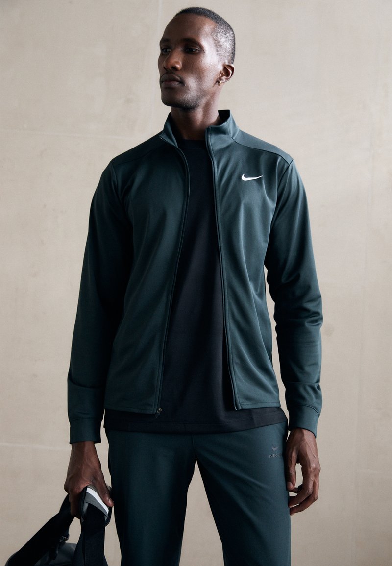 Mørkegrøn atletisk zip-up jakke med høj krave, parret med en sort skjorte og matchende bukser. Har Nike-logoet på jakken.