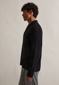 Camisa polo preta de manga longa com um tecido texturizado, colarinho abotoado e um corte relaxado, combinada com calças de moletão cinzas. Vista de perfil lateral.