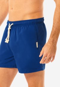 Shorts da bagno blu con vita elastica, chiusura con cordoncino e tasca laterale. Texture liscia; piccola etichetta con logo sul lato.