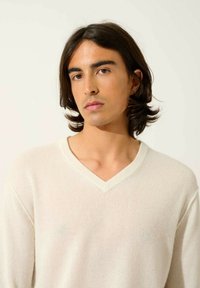 Maglione a V in beige chiaro, realizzato in un tessuto morbido. Presenta una leggera texture e una rifinitura a contrasto lungo il colletto.