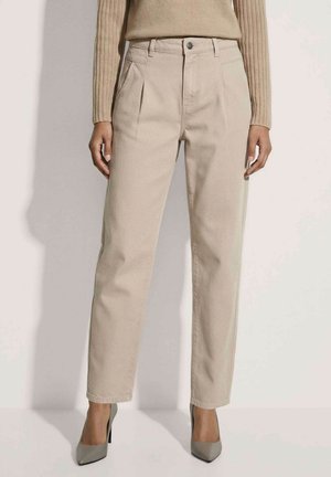 Vrouw draagt beige, rechte hoog taille broek met plooien en grijze puntige pumps, staand tegen een effen lichte achtergrond.
