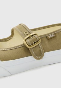 Shoe slip-on de lona bege com sola de borracha branca, apresentando uma fivela de metal dourada, costura branca em contraste e uma etiqueta com o logótipo da Vans.