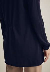 Marinefarbener Langarmtop mit tailliertem Schnitt und lockerem Saum. Glattes Material mit leichtem Glanz, mit subtilen Raffungselementen auf der Rückseite.