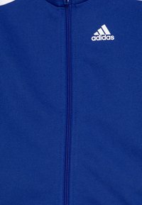 Chaqueta azul con cremallera, fabricada en un tejido suave, que presenta un cuello alto y el logo blanco de adidas en la parte superior izquierda. La cremallera se encuentra en el centro.