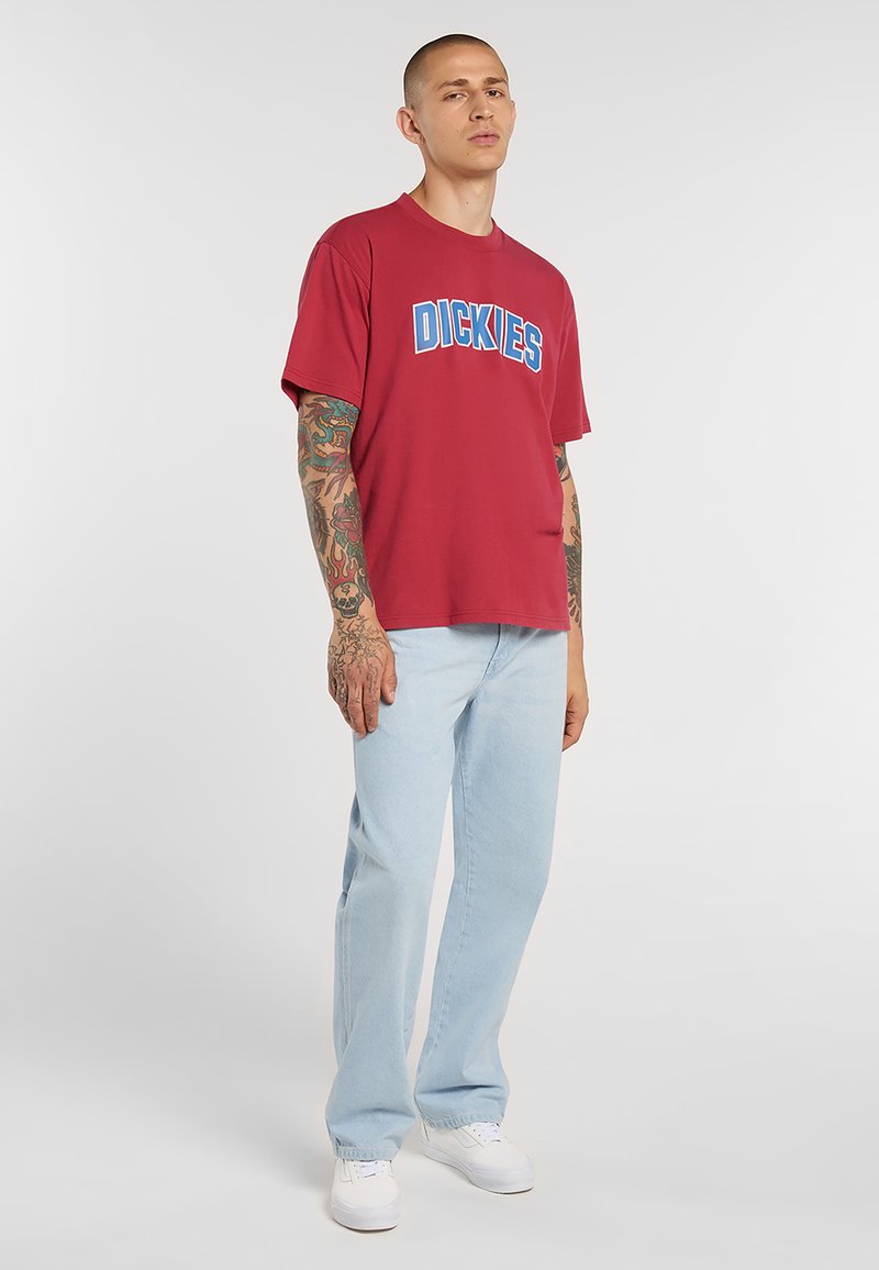 Jonge man met getatoeëerde armen, gekleed in een rood "Dickies" T-shirt, lichtblauwe jeans en witte sneakers, staand tegen een effen achtergrond.