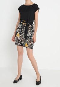 Mujer que lleva un vestido negro de manga corta con una falda de patrón floral y un cinturón negro. Lo complementa con zapatos de tacón negros.