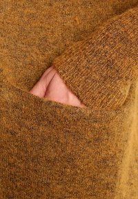 Suéter marrón texturizado con un material suave y de felpa. Presenta un gran bolsillo frontal y puños de punto, mostrando una cálida paleta de colores terrosos.