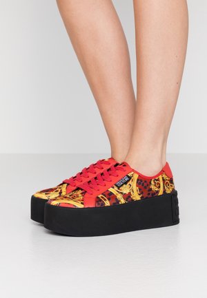 Nogi w czerwono-złotych wzorzystych sneakersach na platformie, z grubymi czarnymi podeszwami, na gładkiej szarej podłodze na tle prostego, jasnego tła.