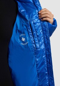 Mėlyna termoizoliacinė striukė su blizgia, siuvinėta vidine dalimi, su logotipu „KHUJO ORIGINAL“ ir šonine kišene.