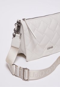 Valkoinen tikattu olkalaukku, jossa on hopeaketju ja säädettävä kangashihna, jossa on kohokuvioitu brändin nimi "Liu Jo."