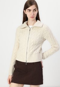 Cardigan creme em malha de cordão com bainha e gola caneladas, apresentando um fecho frontal com zíper. O modelo combina-o com uma saia castanha riscada.
