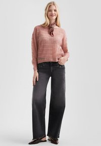Helles rosa gestricktes Sweatshirt mit offenem Muster, kombiniert mit schwarzen weiten Jeans mit Metallösen und dunklen Slipschuhen.