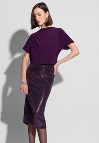 Fioletowa bluzka z drapowaniem w połączeniu z dopasowaną ołówkową spódnicą z fioletowych cekinów. Dodatki obejmują srebrną bransoletkę. Modelka pozuje na tle szarego tła.