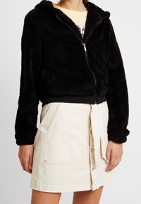 Person som bär en svart zip-up fuzzy jacka och en krämfärgad knappar-för-vidare kjol med stora fickor och "IVY" broderi.