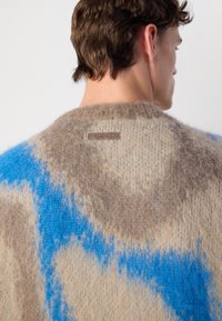 Kötött pulóver bézs és kék árnyalatokban, texturált felülettel. Kerek nyakkivágással rendelkezik, és a hátulján Calvin Klein logós címke található.