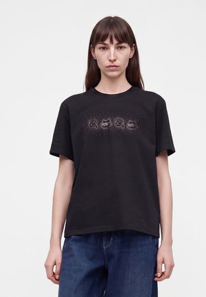 Femme aux cheveux bruns et raides portant un T-shirt noir avec de petits motifs de visage de chat sur la poitrine et un jean bleu.