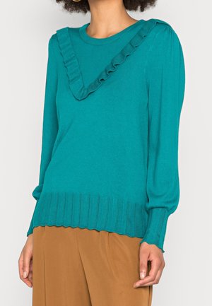 Pull à manches longues turquoise avec un détail volanté en forme de V sur la poitrine, ourlet et poignets côtelés, associé à un pantalon marron.