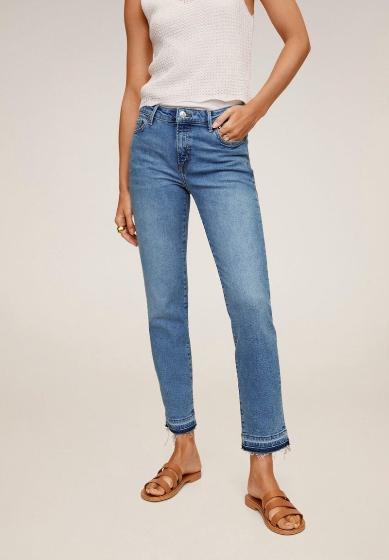Femme portant un jean court bleu clair avec des ourlets effilochés, un top blanc sans manches et des sandales plates marron, debout avec une main dans la poche.