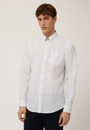White long-sleeve shirt made of smooth fabric. Features button-down collar, front pocket, and subtle logo accent on the pocket.