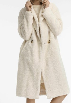 Femme portant un long manteau teddy crème texturé, tenant le col avec les deux mains.
