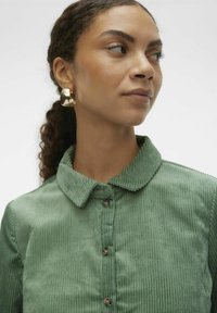 Chemise en velours côtelé vert avec un col, des côtes verticales et un léger éclat. Présente des boutons ronds et un modèle portant des boucles d'oreilles créoles dorées.