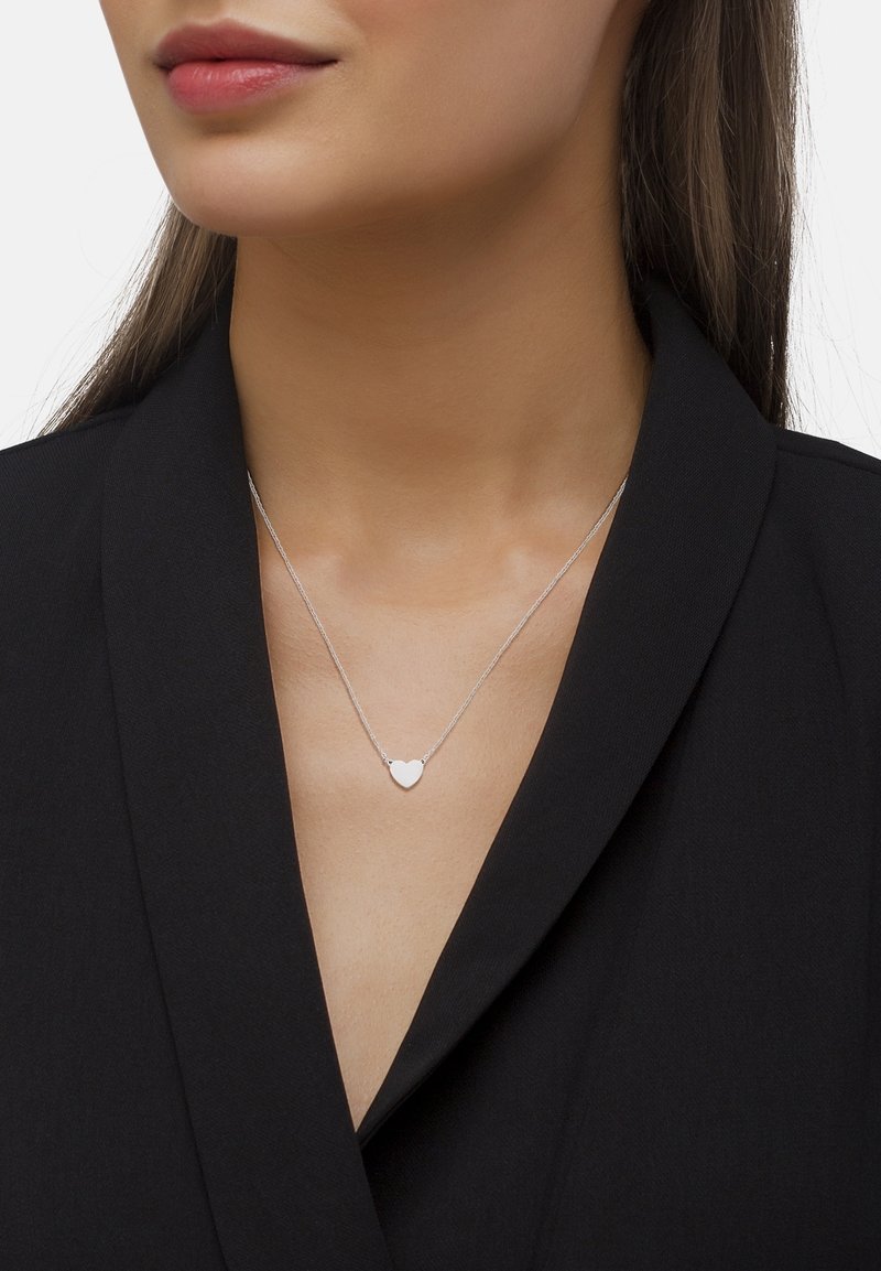 Femme portant un délicat collier pendentif en forme de cœur en argent sur un haut noir à col en V avec de longs cheveux raides.