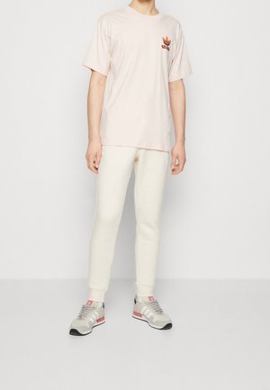 T-shirt en coton rose clair à manches courtes avec un petit logo brodé rouge et noir, associée à un pantalon de survêtement ajusté de couleur crème et des baskets gris.