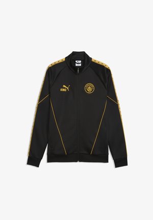 Zwarte jas met volledige rits, gouden bies, Puma-logo en tekst "KING" op de linkerborst, Manchester City-wapen op de rechterborst, en goudkleurige patroonstrepen op de mouwen.