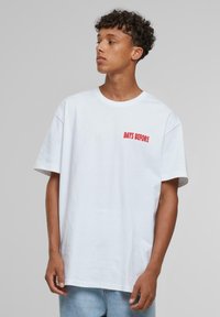 White cotton t-shirt with short sleeves, featuring red embroidered text "DAYS BEFORE" on the left chest area. Casual, loose fit.