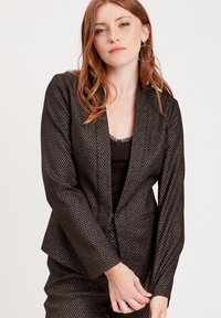 Black blazer with a textured pattern of small metallic accents, featuring a slim fit, lapel collar, and long sleeves. Styled over a black top.