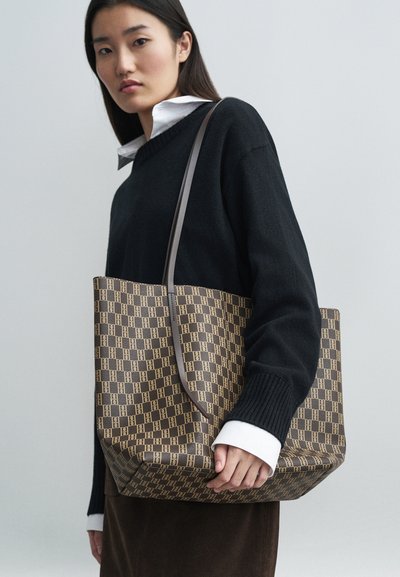 Brązowa torba tote w wzory o strukturalnym kształcie, z gładką teksturą i pojedynczym uchwytem. Model ma na sobie czarny sweter na białej koszuli.