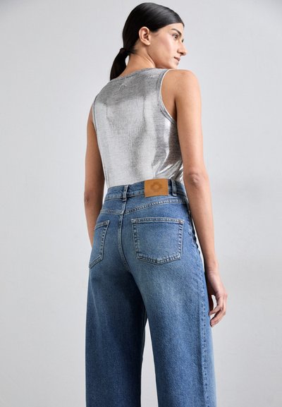 Top sin mangas de color plata metálico combinado con unos jeans de mezclilla azul de pierna ancha y cintura alta, que presentan una parche de cuero marrón en la parte trasera de la cintura.