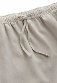 Pantaloni in tessuto beige chiaro con vita elastica e cordoncino annodato al centro davanti, che mostra il materiale lavorato a maglia testurizzato.