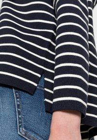 Navy-blau gestreifter Pullover mit weißen horizontalen Streifen, strukturiertem Strickstoff und seitlichen Schlitzen. Kombiniert mit blauen Jeans.