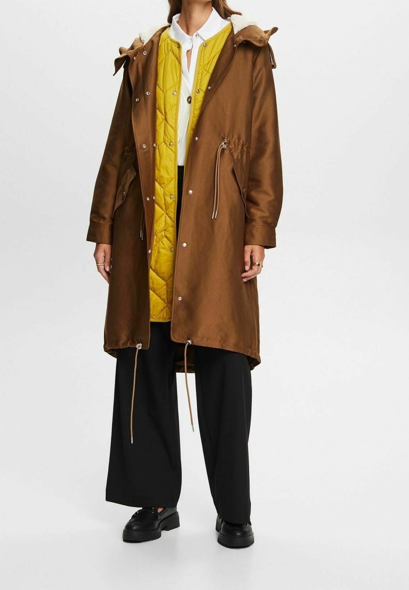 Parka marron imperméable avec une doublure intérieure matelassée jaune, taille à cordon de serrage et bordure en fausse fourrure sur la capuche, associée à un pantalon noir et des chaussures noires.