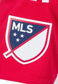 Červená látka s bílým a modrým logem MLS, ve tvaru št ítu s pěti hvězdami, obklopená červeným obrysem a diagonálním pruhovým detailem.