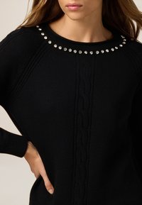 Schwarzer, gestrickter Pullover mit einem strukturierten Zopfmuster, runder Halsauschnitt verziert mit runden Strasssteinen und gerippten Bündchen.