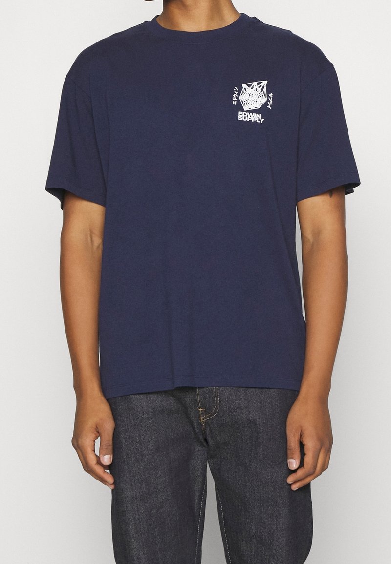 Personne portant un t-shirt bleu marine avec un petit motif géométrique blanc et le texte « EDWIN SUPPLY » sur la poitrine gauche, associé à un jean foncé.