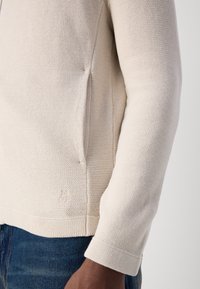 Lys beige strikkegenser med en strukturert finish. Har en sidelommepocket og diskret merke ved kanten. Avslappet passform med lange ermer.